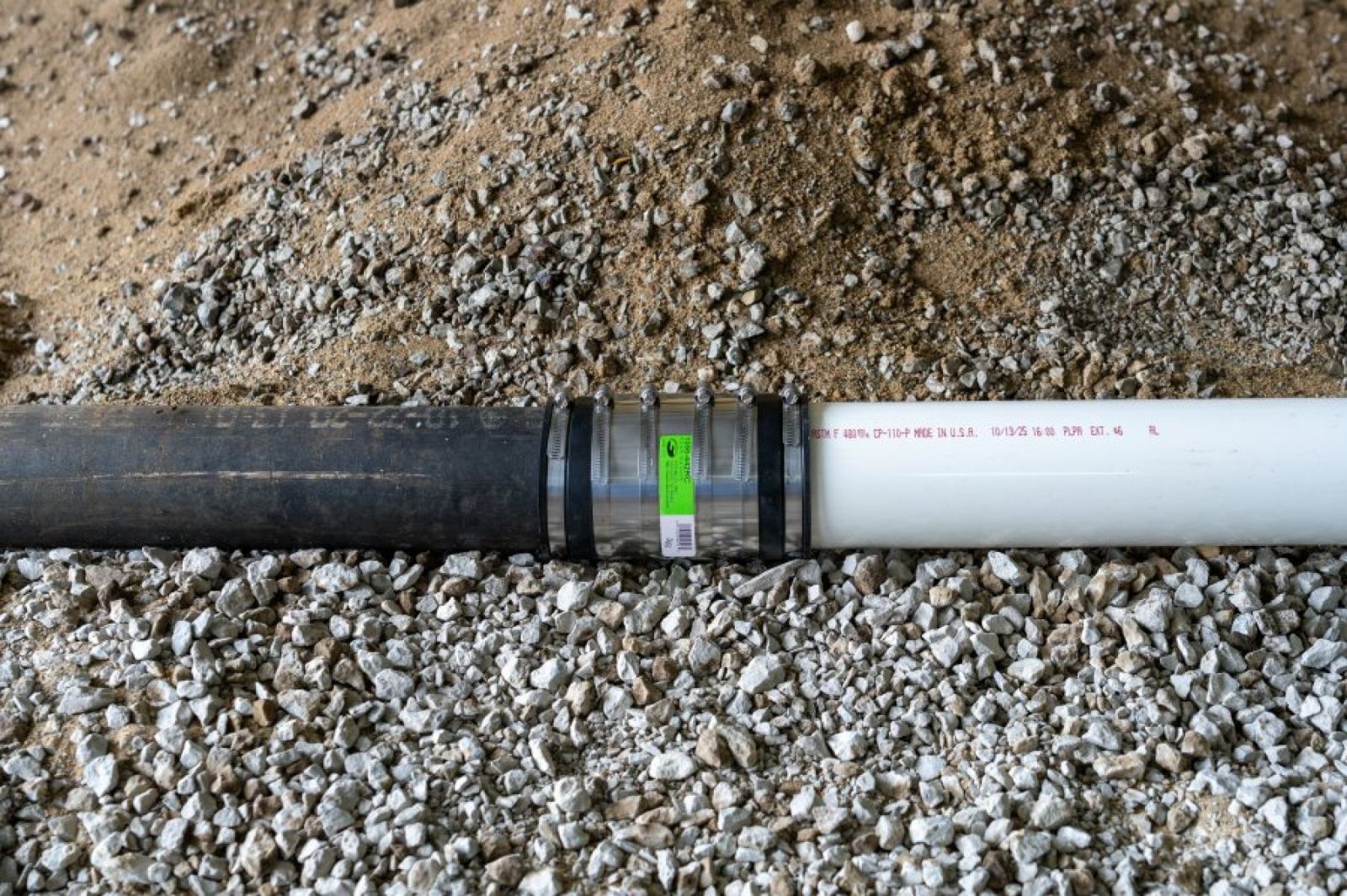 Fernco XL coupling joining black pipe to white PVC pipe on a gravel bed